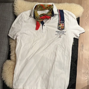 Vit pikétröja från Aeronautica Militare - Säljer en snygg vit pikétröja från Aeronautica Militare med coola detaljer som broderade emblem och ränder i blått och rött. Perfekt för en stilren och sportig look. Tröjan har korta ärmar och en klassisk krage. Passar både till vardags och fest!