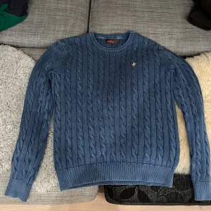 Blå kabelstickad tröja från Morris - Säljer en snygg blå kabelstickad tröja från Morris. Den har en klassisk design med rund hals och ett litet broderat emblem på bröstet. Perfekt för kyliga dagar och passar både till vardags och lite finare tillfällen. 🧶💙