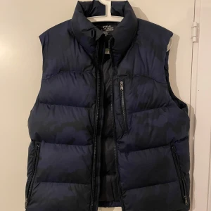 Blå dunväst från Polo Ralph Lauren - Säljer en snygg blå dunväst från Polo Ralph Lauren. Västen har en dragkedja framtill och två praktiska fickor med dragkedjor. Perfekt för kyliga dagar och ger en stilren look. Passar bra till både vardag och lite finare tillfällen.