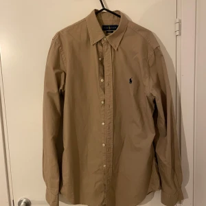 Beige skjorta från Ralph Lauren - Säljer en snygg beige skjorta från Ralph Lauren i slim fit. Skjortan har långa ärmar och klassiska knappar framtill. Perfekt för både vardag och festliga tillfällen. Den är i mycket bra skick och redo för en ny ägare!