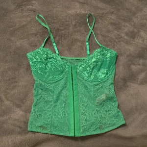 Grön spetskorsett från NA-KD - Snygg grön korsett från NA-KD i spetsmaterial. Den har justerbara axelband. Perfekt för en utgång. Korsetten är aldrig använd, alltså nyskick! 