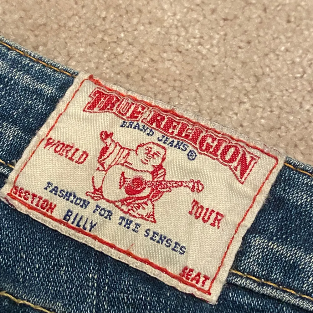 Säljer ett par snygga blå jeans från True Religion i ny skick. De har en tight passform och är lågmidjade med klassiska femfickor och knappar bak. Perfekta för både vardag och fest! 🕺. Jeans & Byxor.