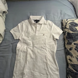 Vit pikétröja från Tommy Hilfiger - Säljer en klassisk vit pikétröja från Tommy Hilfiger i storlek XS. Tröjan har en slim fit passform och är tillverkad i ett stretchigt material för extra komfort. Perfekt för både vardag och lite mer uppklädda tillfällen. Den har korta ärmar och en liten logga på bröstet. Oanvänd!