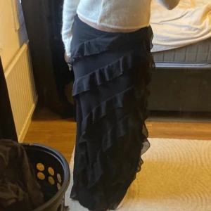 Svart långkjol med volanger - Säljer en elegant svart långkjol med volanger. Kjolen har en fin passform och är perfekt för både fest och vardag. Materialet känns mjukt och bekvämt, vilket gör den idealisk för höst och vinter. Passar bra med en stickad tröja eller blus. Perfekt för att skapa en chic look!