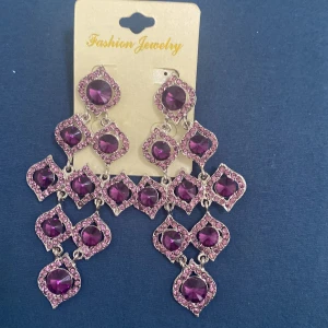 Lila glittriga örhängen - Säljer ett par fantastiska lila örhängen från Fashion Jewelry. De är stora och glittriga med flera lila stenar i en vacker design. Perfekta för att lysa upp vilken outfit som helst på festen!