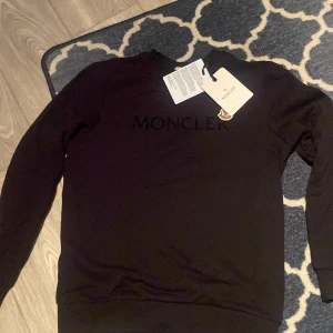 Svart tröja från Moncler - Säljer en snygg svart tröja från Moncler. Den är i nyskick och har en stilren design med Moncler-loggan på bröstet. Tröjan har långa ärmar och är perfekt för både vardag och fest. Materialet känns mjukt och bekvämt.