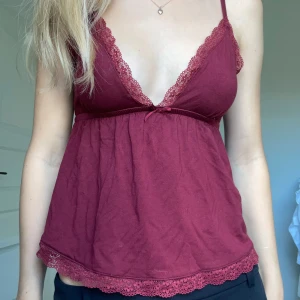 Vinröd linne med spetsdetaljer - Säljer en superfin vinröd linne med spetsdetaljer. Den har en v-ringad design och en söt liten rosett framtill. Perfekt för sommardagar eller en mysig hemmakväll. Materialet känns mjukt och bekvämt mot huden. Från nelly storlek XS