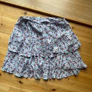 Blommig kjol från Zara - Säljer en superfin blommig kjol från Zara i storlek XS. Kjolen är ljusblå med ett vackert blommönster i rosa och lila nyanser. Den har en volangdetalj som ger den en extra söt touch. Perfekt för vår och sommar!