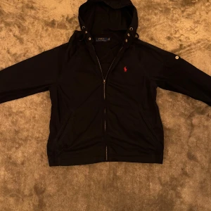  jacka från Polo Ralph Lauren - Marinblå jeansjacka med huva från Polo Ralph Lauren. Jackan har dragkedja framtill, knappdetaljer vid huvan och en röd broderad logga på bröstet. Praktiska fickor och knapp på ärmen ger extra stilpoäng. Inte mycket använd. Nypris ligger på runt 4000kr