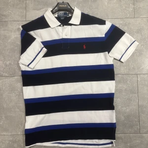 Randig pikétröja från Polo Ralph Lauren - Säljer en klassisk pikétröja från Polo Ralph Lauren i vitt, svart och blått med breda ränder och röd broderad logga på bröstet. Tröjan har korta ärmar och krage med knappar. Perfekt för en sportig och stilren look.