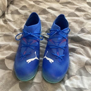 Blåa fotbollsskor från Puma - Säljer ett par blåa fotbollsskor från Puma med vita detaljer och logga på sidan. Skorna har snörning och en smidig, låg profil med platt sula och rund tå. Perfekta för dig som vill sticka ut på planen!