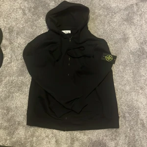Svart zip up Stone Island - Säljer en svart zip up från Stone Island med dragkedja och klassisk logotyp-patch på ärmen. Jackan har huva och en stilren, enkel design som passar perfekt till streetwear-stilen.