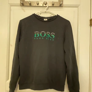 Svart sweatshirt från Hugo Boss - Svart sweatshirt från Hugo Boss med tryckt logga i grönt och vitt på bröstet. Klassisk rund halsringning och långa ärmar. Perfekt för en stilren och avslappnad look. Har en målat fläck som visas på sista bilden. 