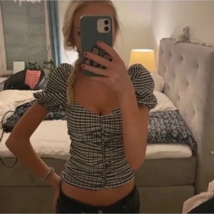 Svartvit rutig blus med puffärmar - Säljer en svartvit rutig blus med puffiga korta ärmar och knappar framtill. Blusen är croppad och har en söt, figurnära passform med lätt rynkade detaljer längs knappraden. Perfekt för dig som gillar trendiga toppar!