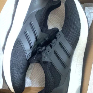 Svarta Adidas Ultraboost sneakers - Svarta Adidas Ultraboost sneakers med stickad ovandel och klassiska tre ränder på sidan. Skorna har vit, tjock och bekväm sula samt snörning framtill. Perfekta för både träning och vardag.