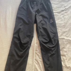 Svarta skalbyxor från Haglöfs - Svarta skalbyxor från Haglöfs med elastisk midja och diskret logga på låret. Lätta och smidiga byxor som passar perfekt för utomhusaktiviteter. Tillverkade i syntetmaterial med meshfoder upptill för extra komfort.