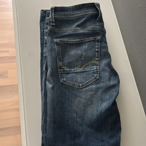 Blå jeans från Jack & Jones - Säljer ett par blå jeans från Jack & Jones med slitna detaljer och knappar i gylfen. Jeansen har klassisk femficksdesign och en något tvättad look med slitningar på låren och knäna.