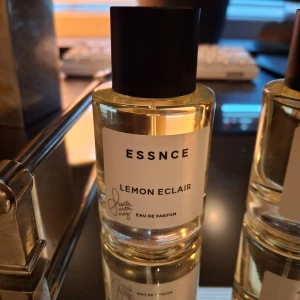 ESSNCE Lemon Eclair Eau de Parfum - Fräsch parfym med doft av citron och sötma, Lemon Eclair från ESSNCE. Flaskan är genomskinlig med svart lock och vit etikett. Perfekt för dig som gillar fruktiga och fräscha dofter. Oöppnad.