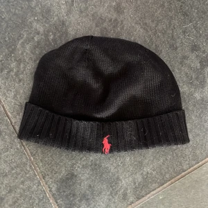 Mössa från Polo Ralph Lauren - Svart stickad mössa från Polo Ralph Lauren med uppvikt kant och röd broderad logotyp framtill. Tillverkad i 100% ull för extra värme och komfort. Klassisk och stilren design som passar till många olika outfits. Lite använd men märks inget bara på insidan