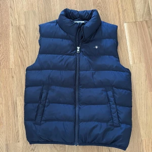 Mörkblå dunväst från GANT - Säljer en mörkblå dunväst från GANT dragkedja framtill. Västen har två fickor på sidorna och klassisk GANT-logga på bröstet. Perfekt för lager-på-lager och stilren look. Använd av ett barn ett fåtal gånger och är i mycket fint skick!