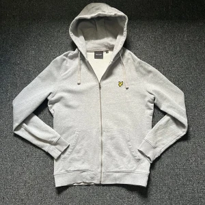 Ljusgrå hoodie Lyle & Scott - Säljer en ljusgrå hoodie från Lyle & Scott med dragkedja och huva. Tröjan har två fickor framtill och den klassiska gula loggan på bröstet. Perfekt för en avslappnad stil. Är i Storlek S och är i mycket bra skick med inga defekter på. Hör av er vid frågor eller funderingar!