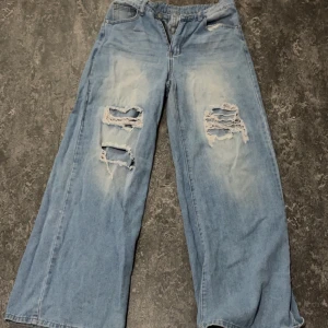 Ljusblå wide leg jeans med slitningar - Säljer ett par ljusblå jeans med breda ben och stora slitningar på båda benen framtill. Jeansen har klassisk femficksdesign och knappgylf. Perfekta för en avslappnad och trendig look. 