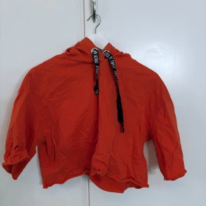 Orange croppad hoodie med tryckta snören - Säljer en orange croppad hoodie med svarta snören där det står 'DIVIDED'. Hoodien har långa ärmar och en avslappnad passform, perfekt för en cool och avslappnad stil.