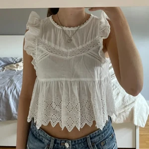 Vit blus med spets och volang från Zara - Säljer en söt vit blus från Zara med spetsdetaljer och volang vid axlarna. Blusen är croppad och har ett broderat mönster nertill samt vågig kant. Storlek xs men passar även s👼🏼👼🏼 Knappen bak har lossnat, men är nog superenkelt att sy på ny om så önskas (Men det behövs ej). 