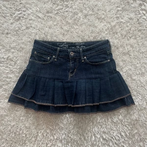 Mörkblå jeanskjol med volang - En lågmidjad jeanskjol omsydd från ett par jeans. Midjemått tvärsöver - ca 35cm Längd - 32cm