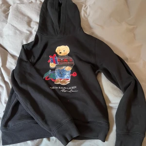 Svart hoodie med björn från Polo Ralph Lauren - Svart hoodie från Polo Ralph Lauren med ikonisk Polo Bear-tryck på framsidan. Björnen har jultröja och håller i paket, vilket ger en festlig känsla. Tröjan har huva och är tillverkad i mjukt material.
