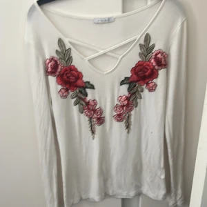 Vit långärmad topp med broderade blommor - Säljer en vit långärmad topp från Zara med snygga broderade röda och rosa blommor framtill. Toppen har v-ringning med korsade band och är i mjukt material. Perfekt för dig som vill ha något unikt och trendigt i garderoben!
