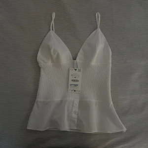 Vit topp från Zara  - En helt ny och snygg vit topp med smala axelband och v-ringning. Toppen har en ribbad mittdel och knappar framtill. Ny pris 329kr