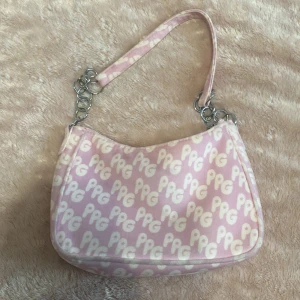 Rosa axelväska med PG-mönster - Säljer en söt rosa axelväska med vitt PG-tryck över hela väskan. Väskan har silverfärgade kedjedetaljer på axelremmen och stängs med dragkedja. Insidan är fodrad i ljusrosa tyg. Perfekt för att addera lite färg till din outfit!