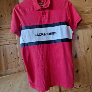 Röd piké t-shirt från Jack & Jones - Röd piké t-shirt från Jack & Jones med vit och svart rand över bröstet och tryckt logga. Klassisk krage med knappar framtill. Perfekt för en avslappnad och sportig stil.