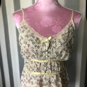 Blommig linne med spets från Vila - Söt ljus klänning från Vila med smala axelband, spetsdetaljer och små blommor i brunt. Klänningen har en liten rosett framtill, knappar och markerad midja med gul kant. Perfekt för varma dagar.storlek M på lappen men mindre som S