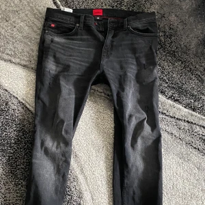 Svarta slim jeans från Hugo - Säljer ett par svarta slim fit jeans från Hugo med diskret slitning på framsidan. Jeansen har klassisk femficksdesign, dragkedja och knapp i midjan. Snygg tvätt med lite ljusare partier och röda detaljer på insidan av linningen. Priset går såklart att diskuteras