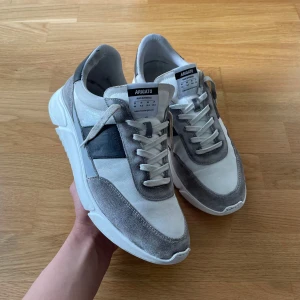 Grå och vita sneakers från Axel Arigato - Snygga sneakers från Axel Arigato i vitt och olika nyanser av grått med svarta detaljer. Skorna har snörning, rund tå och en platt sula. Ovandelen är i mocka och syntetmaterial, vilket ger en modern och stilren look.