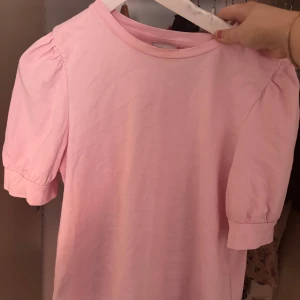 Ljusrosa topp från Aware by Vero Moda - Säljer en ljusrosa topp från Aware by Vero Moda. Toppen har korta puffärmar och rund halsringning. Tillverkad i mjukt bomullsmaterial, perfekt för vår och sommar. Enkel och stilren design som passar till mycket.