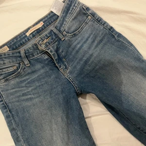 Levis 501 blå jeans - Klassiska blå Levis 501 jeans med raka ben och fem fickor. Byxorna har normal passform, knappgylf och tydliga Levis-detaljer som läderpatch och röd etikett vid bakfickan. Perfekta för en avslappnad stil.