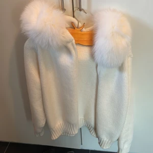 Vit stickad jacka med päls på luvan - Mysig vit stickad jacka med stor huva och fluffig päls runt huvan från Alessa, aldrig använt!!. Jackan har ribbade muddar och en avslappnad passform.