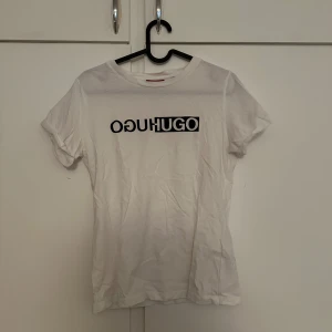 Vit t-shirt med tryck från HUGO - Vit t-shirt från HUGO med svart och vitt logotyp-tryck på bröstet. Klassisk rund halsringning och korta ärmar. Perfekt för en clean och stilren look.