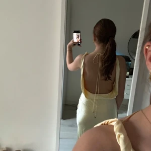 Ljusgul långklänning - Säljer en ljusgul långklänning i satin med smala axelband och snygg öppen rygg med knyt. Klänningen har en elegant omlott-effekt framtill och ett mjukt, glansigt material som faller fint. Passar jätte bra till balen eller ett bröllop. Har aldrig använt klänningen så den är jätte bra skick💛💛!!