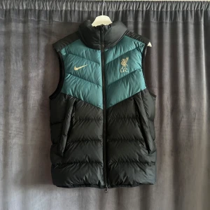 Svart och grön dunväst från Nike x Liverpool FC - Snygg dunväst från Nike i samarbete med Liverpool FC. Västen är svart med gröna partier upptill, dragkedja framtill och två sidofickor. Liverpool FC-logga och Nike-logga i grått på bröstet. Perfekt för kyliga dagar.