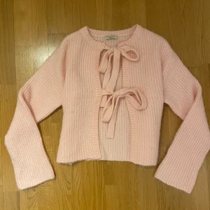 Ljusrosa stickad kofta med rosetter från Gina Tricot - Superfin ljusrosa stickad kofta från Gina Tricot. Koftan är croppad och har två dekorativa rosetter framtill. Mjukt material och långa ärmar, perfekt för lager-på-lager-stil.