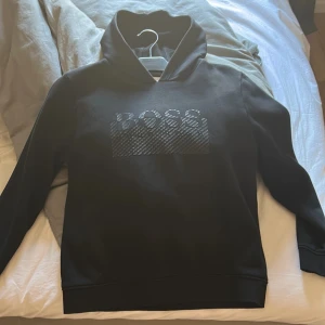 Svart hoodie från Hugo Boss - Säljer en stilren svart hoodie från Hugo Boss med logotypen i ett diskret mönster på bröstet. Hoodien har långa ärmar och en klassisk huva. Priset kan diskuteras.