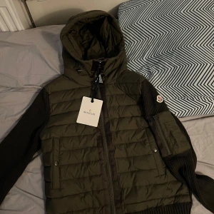 Olivgrön Moncler Cardigan - Säljer en olivgrön Moncler-jacka med quiltad front och huva. Ärmar och rygg är stickade i mörkare nyans, vilket ger en snygg kontrast. Jackan har dragkedja framtill, två fickor med tryckknappar och Moncler-logga på ärmen. Sjukt snygga. 
