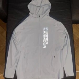 Grå vindjacka med huva från Under Armour - Säljer en grå vindjacka från Under Armour med huva och hel dragkedja. Jackan har svarta detaljer längs ärmarna och två fickor med dragkedja. Stor logga och text på bröstet. 