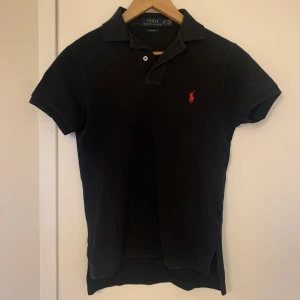 Svart pikétröja från Polo Ralph Lauren - Klassisk svart pikétröja från Polo Ralph Lauren med röd broderad logga på bröstet. Tröjan har korta ärmar, krage och två knappar vid halsen. Perfekt för en stilren och avslappnad look. Storleken är XS och jag skulle säga att den passar någon mellan runt 165-170 och som väger runt 55kg. Skicket är ca 7/10