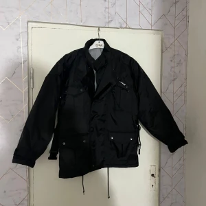 Svart jacka från Spruce - Säljer en svart jacka från Spruce med flera fickor framtill och tryckknappar. Jackan har hög krage, dragsko i midjan och lång ärm. Perfekt för dig som gillar en stilren och funktionell look.