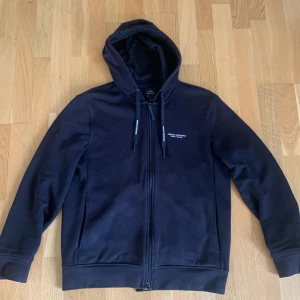 Svart hoodie från Armani Exchange - Säljer en svart hoodie från Armani Exchange i storlek M. Tröjan har dragkedja framtill, huva med snören och diskret logga på bröstet samt på snörena. Perfekt för en stilren och avslappnad look. Pris på nätet 1800 kr.